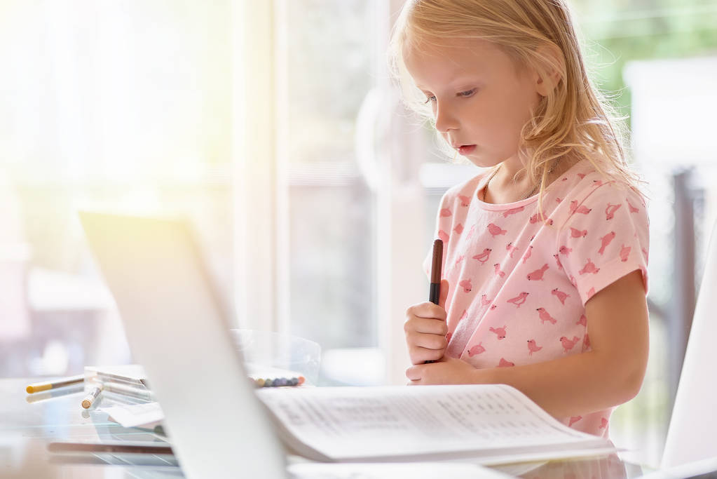 Pieni tyttö vaaleanpunaisessa t-paidassa tekee läksynsä kannettavalla. Valoisa huone päivänvalossa, itsenäinen tyttö, koulutus
 - Valokuva, kuva