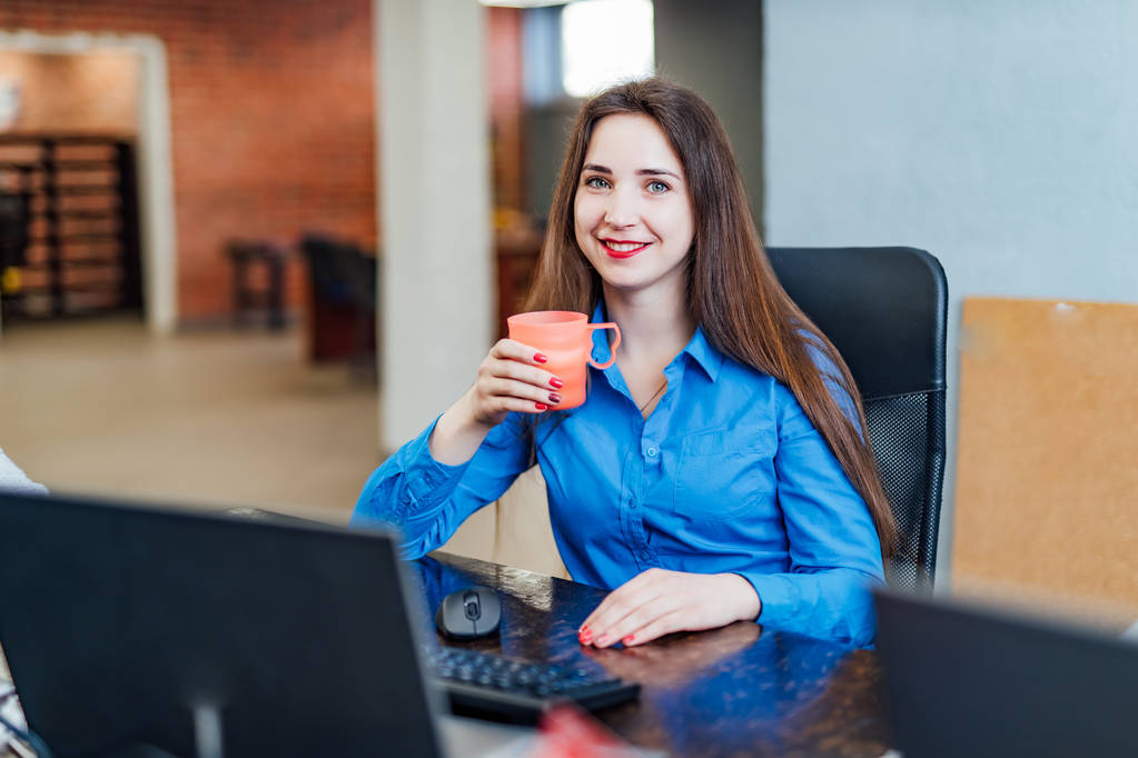 Jovem programadora sentada em um escritório da empresa em frente ao computador com uma xícara rosa. Engenheiro de informática profissional olhando para a câmera e sorrindo. Trabalho de software
 - Foto, Imagem
