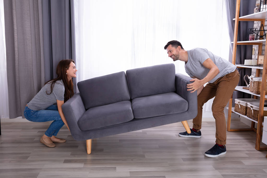 Vista lateral de una sonriente pareja joven que levanta el sofá en la sala de estar
 - Foto, Imagen