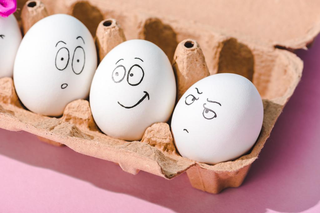 eggs with surprised, angry and smiling face expressions in egg carton on pink - Photo, Image