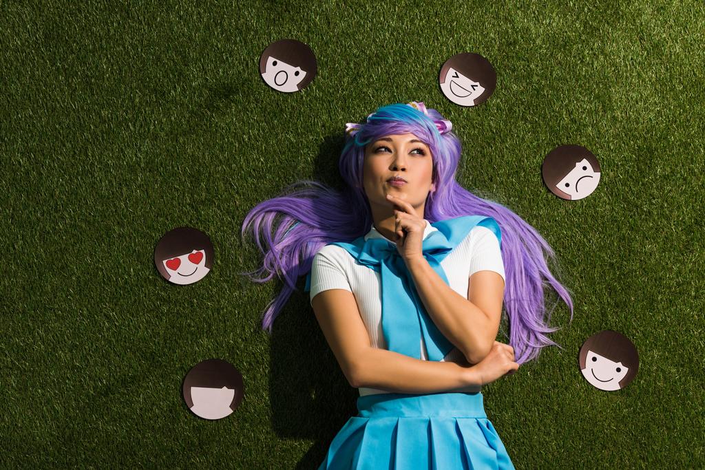 nachdenkliches asiatisches Anime-Mädchen in lila Perücke mit Emoticons auf Gras liegend - Foto, Bild