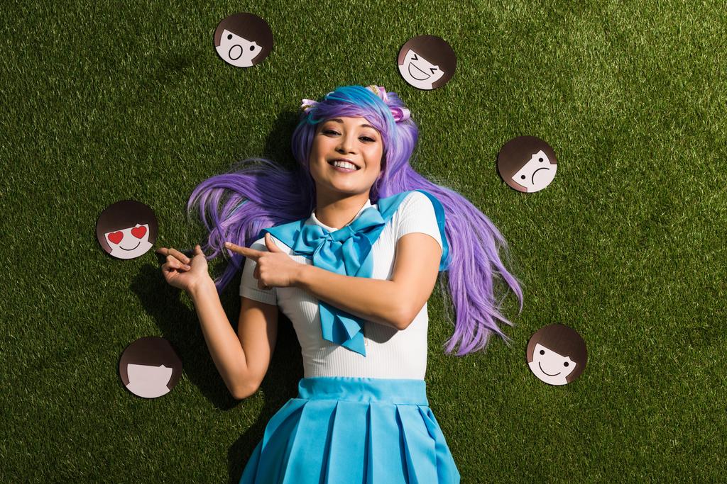 Aziatische anime meisje in paarse pruik met emoticons liggend op gras - Foto, afbeelding