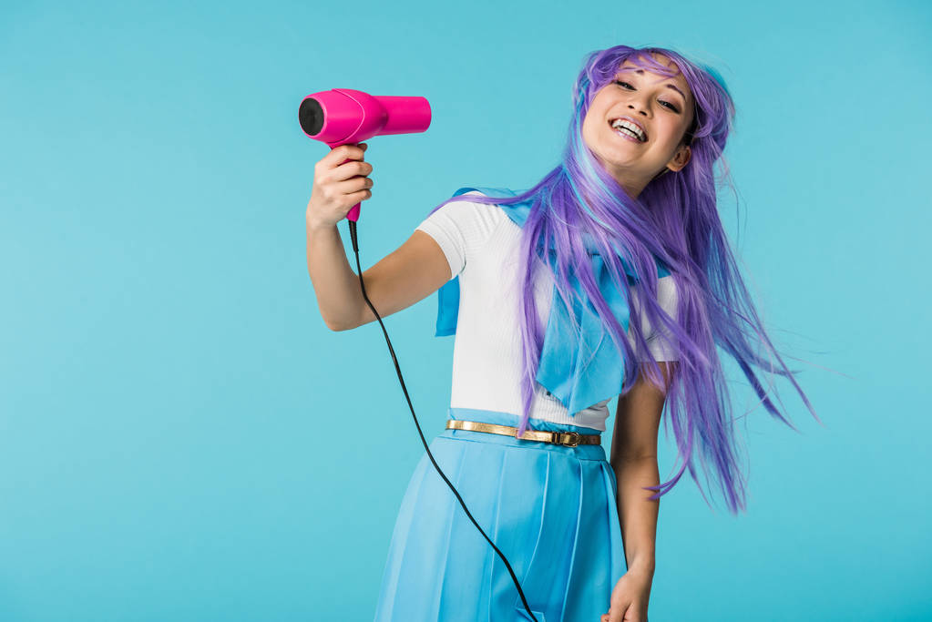 Smiling asian anime girl in wig using hairdryer isolated on blue - Photo, Image