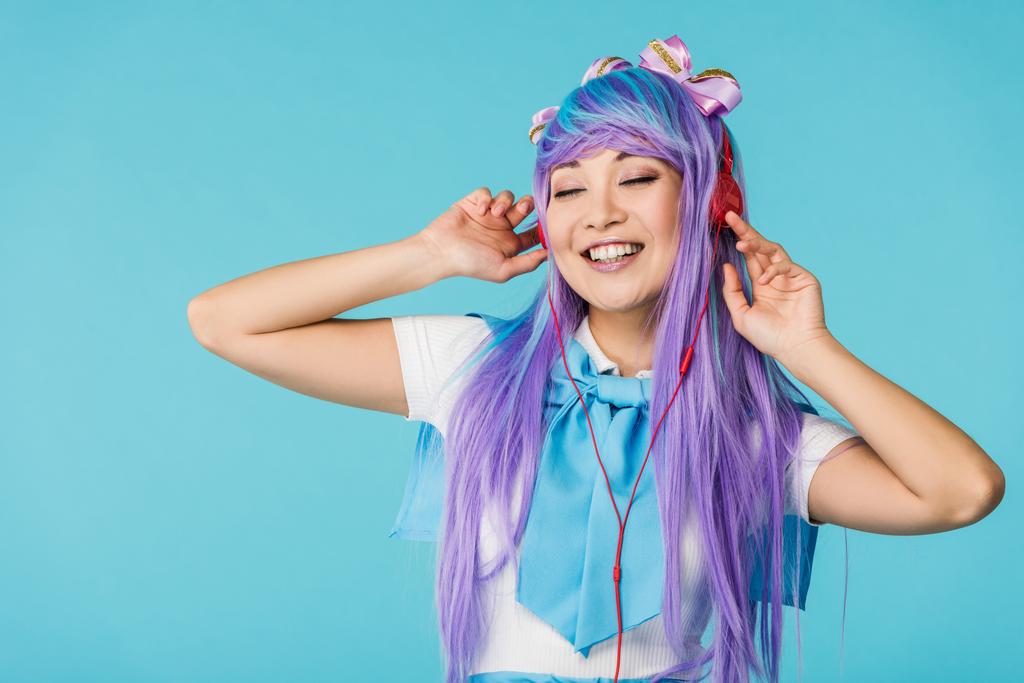 Χαμογελαστός κορίτσι anime σε μωβ περούκα ακούγοντας μουσική στα ακουστικά που απομονώνονται στο μπλε - Φωτογραφία, εικόνα