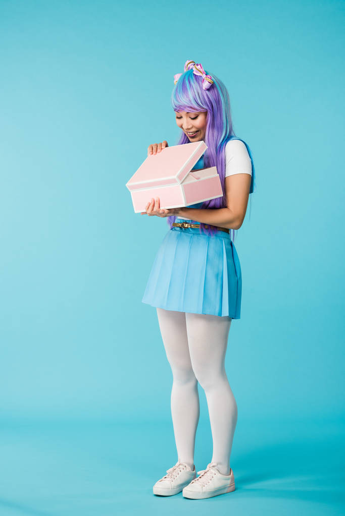 Вид в полный рост на живую девушку в фиолетовом парике, держащую в руках кейс на голубом фоне
 - Фото, изображение