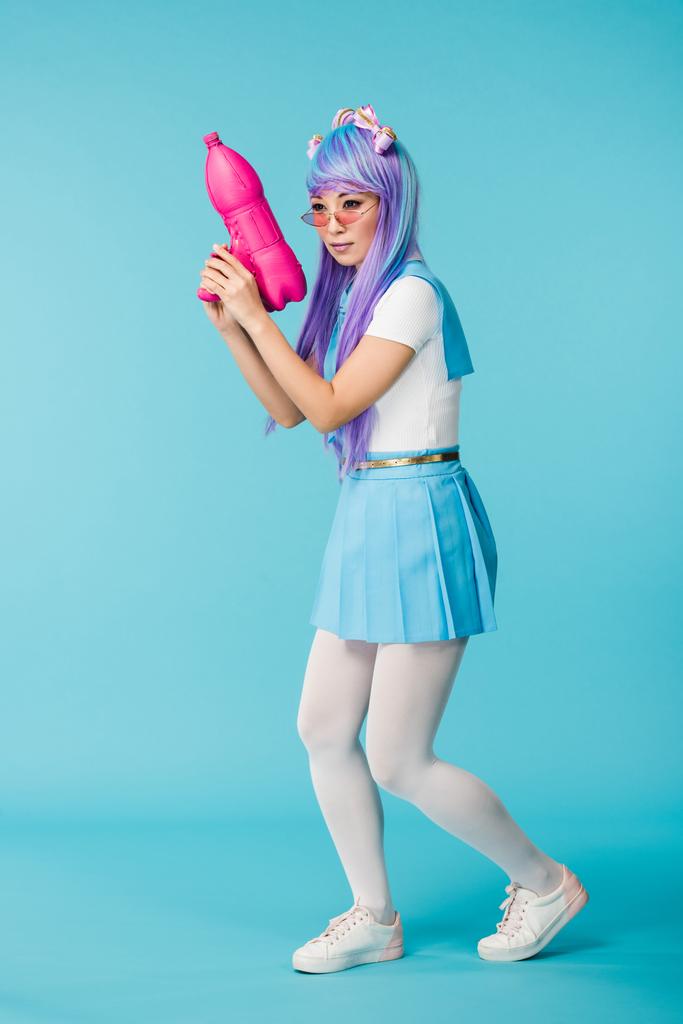 Plná délka pohledu na asijskou anime dívku v paruce a brýlích s vodní pistolí na modrém - Fotografie, Obrázek