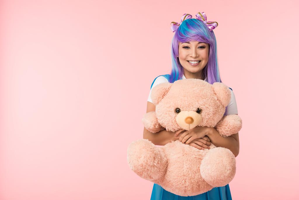 Χαμογελαστός ασιατικό κορίτσι anime σε περούκα κρατώντας αρκουδάκι απομονωθεί σε ροζ - Φωτογραφία, εικόνα