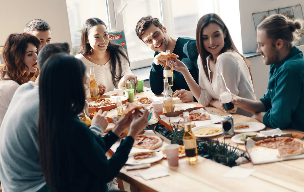 Grupo de jóvenes en ropa casual comiendo pizza y sonriendo mientras hacen una cena en el interior
                - Foto, Imagen
