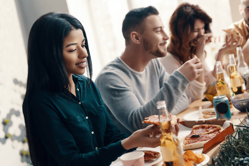 unter besten Freunden. Gruppe junger Leute in Freizeitkleidung isst Pizza und lächelt, während sie drinnen eine Dinnerparty feiern - Foto, Bild