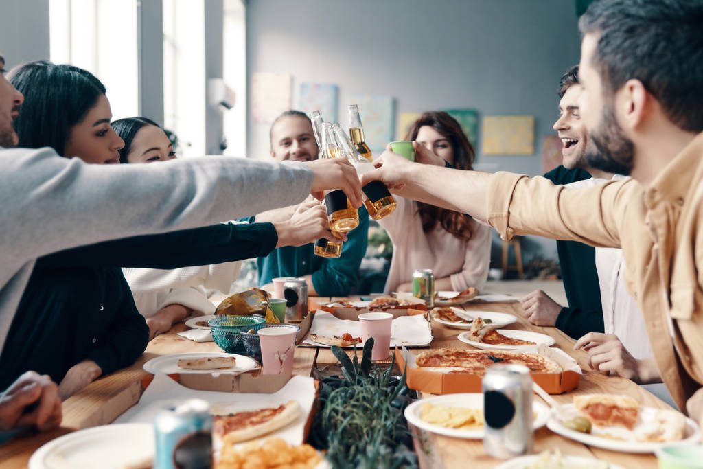 Şerefe! Gündelik giyimli bir grup genç, içeride akşam yemeği partisi verirken birbirlerine kadeh kaldırıyor ve gülümsüyor          - Fotoğraf, Görsel