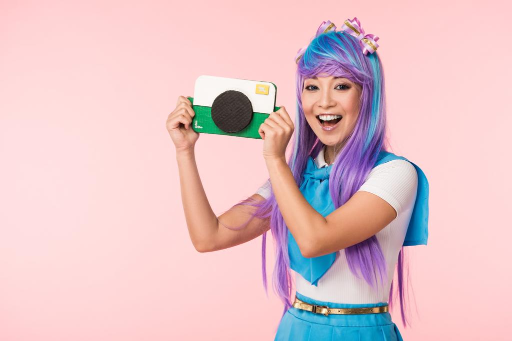 Sonriente asiático anime chica en púrpura peluca celebración de cartón cámara aislada en rosa
 - Foto, imagen