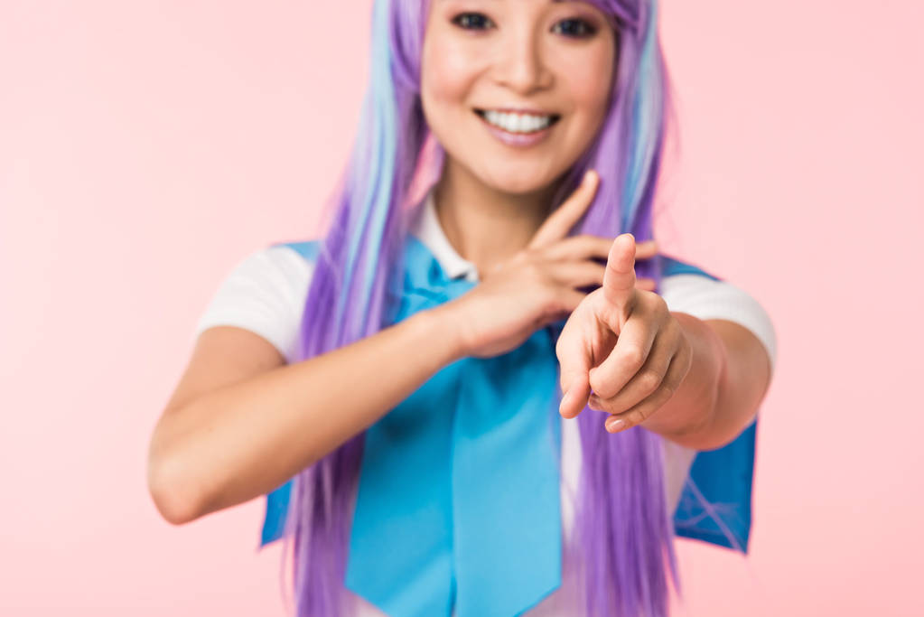 lächelnde asiatische Anime-Mädchen in lila Perücke zeigt mit dem Finger auf Kamera isoliert auf rosa - Foto, Bild