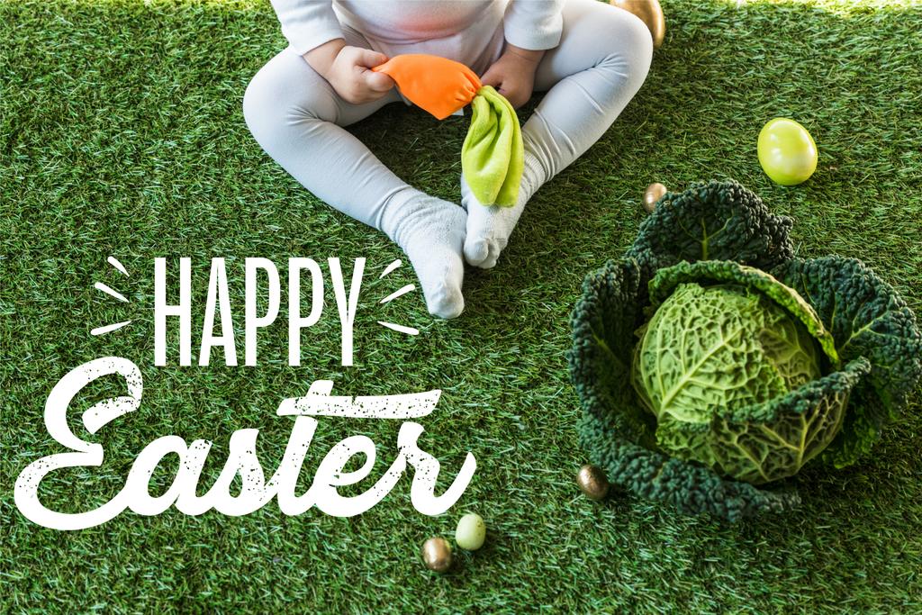 visão parcial da criança segurando cenoura de brinquedo enquanto sentado perto de ovos de Páscoa e couve lombarda na grama verde com letras felizes da Páscoa
 - Foto, Imagem