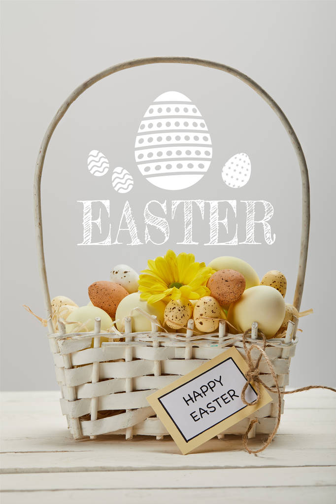 wicker basket with colorful eggs, yellow gerbera flower, greeting card and Easter lettering on grey background - Photo, Image