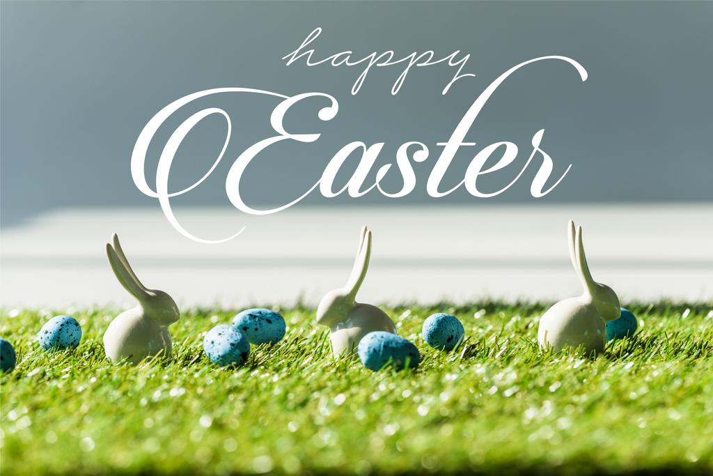 decorative rabbits on green grass near blue quail eggs with happy Easter lettering - Photo, Image