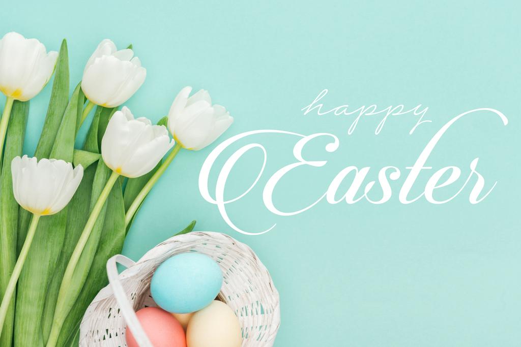 Top uitzicht van geschilderde kip eieren in rieten mand en witte tulpen op blauwe achtergrond met witte Happy Easter belettering  - Foto, afbeelding