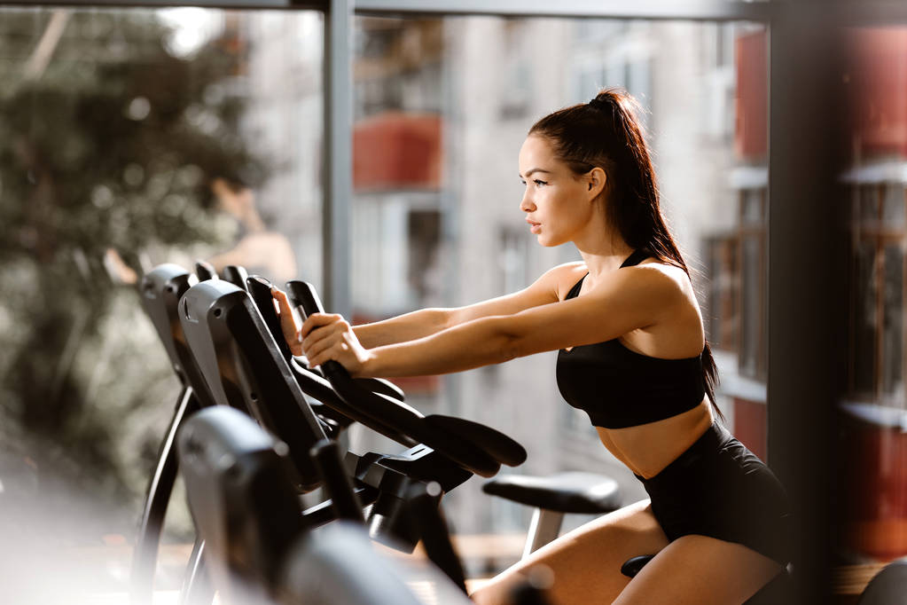Νεαρή αθλητική κοπέλα με καστανά μαλλιά ντυμένη με μαύρο αθλητικό μπλουζάκι και σορτς γυμνάζεται στο ποδήλατο γυμναστικής στο γυμναστήριο - Φωτογραφία, εικόνα
