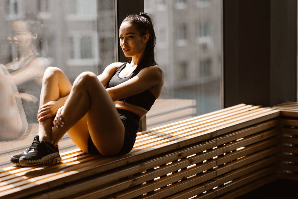 Athletic sötét hajú lány öltözött, fekete sportok felső és rövidnadrág ül egy fából készült ablakpárkányon az edzőteremben - Fotó, kép