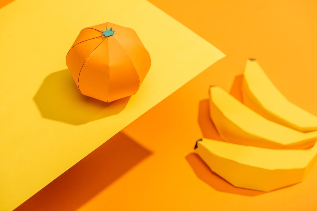 Selektivní zaměření origami mandarinky na žlutém papíře s lepenkovou banány na oranžovém - Fotografie, Obrázek
