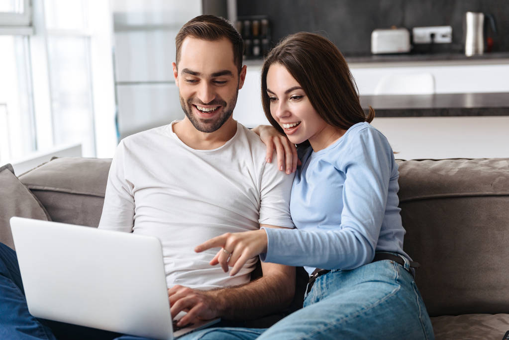 Imagen de pareja feliz usando el ordenador portátil juntos mientras están sentados en sof
 - Foto, Imagen