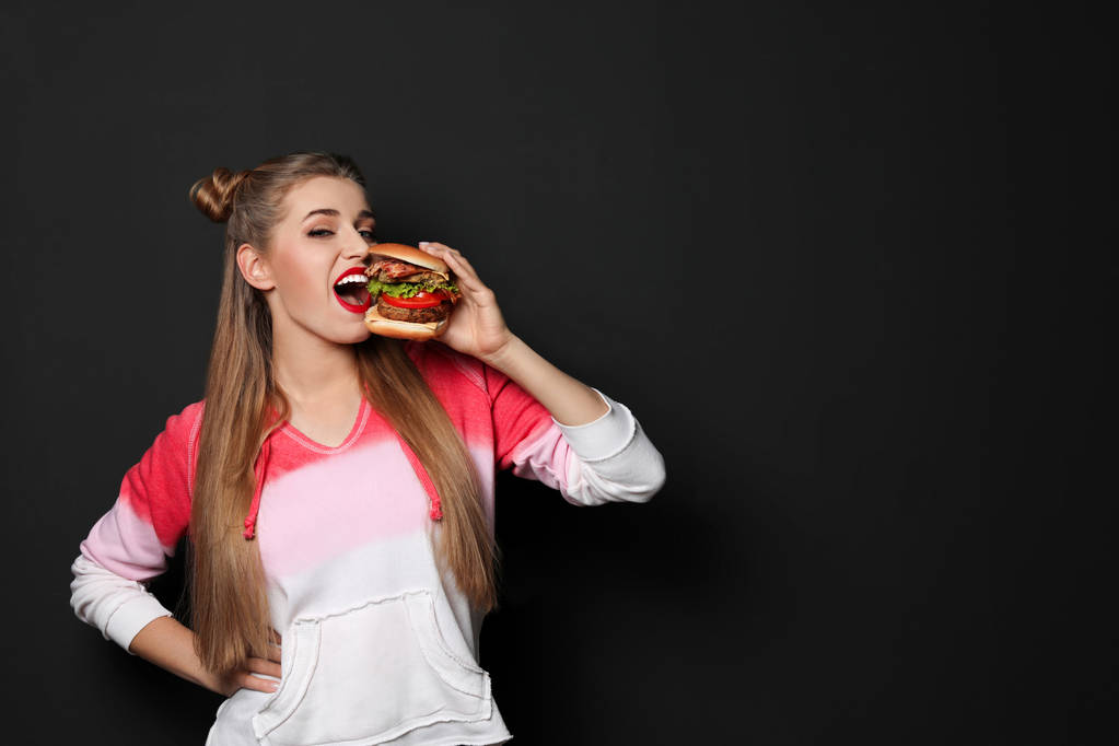 hübsche Frau isst leckeren Burger auf schwarzem Hintergrund. Raum für Text - Foto, Bild