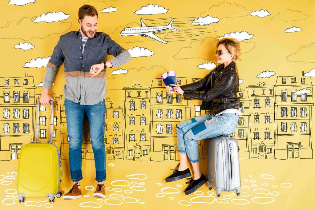 黄色の背景に都市と飛行機のイラストでスーツケースを持つ男性が時間をチェックしながら、パスポートとチケットを保持している幸せな女性 - 写真・画像