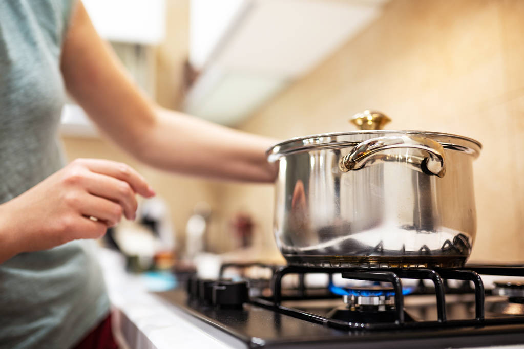 Красивая молодая женщина домохозяйка готовит ужин, держит в руках большую стальную кастрюлю, стоя на газовой плите
. - Фото, изображение