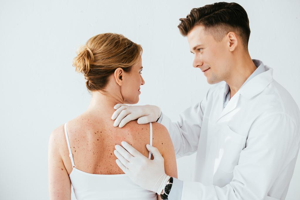 όμορφος δερματολόγος σε γάντια λατέξ εξέταση γυναίκα με μελάνωμα απομονωμένη σε λευκό  - Φωτογραφία, εικόνα