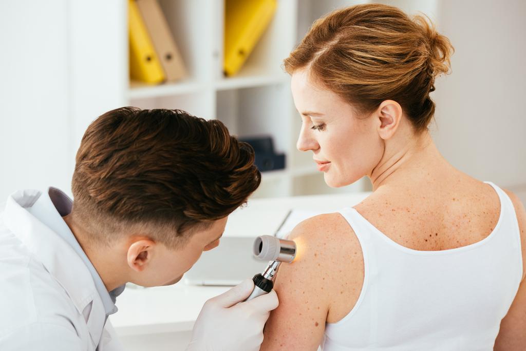 dermatologista em luvas de látex segurando dermatoscópio enquanto examina paciente atraente com doença de pele
   - Foto, Imagem