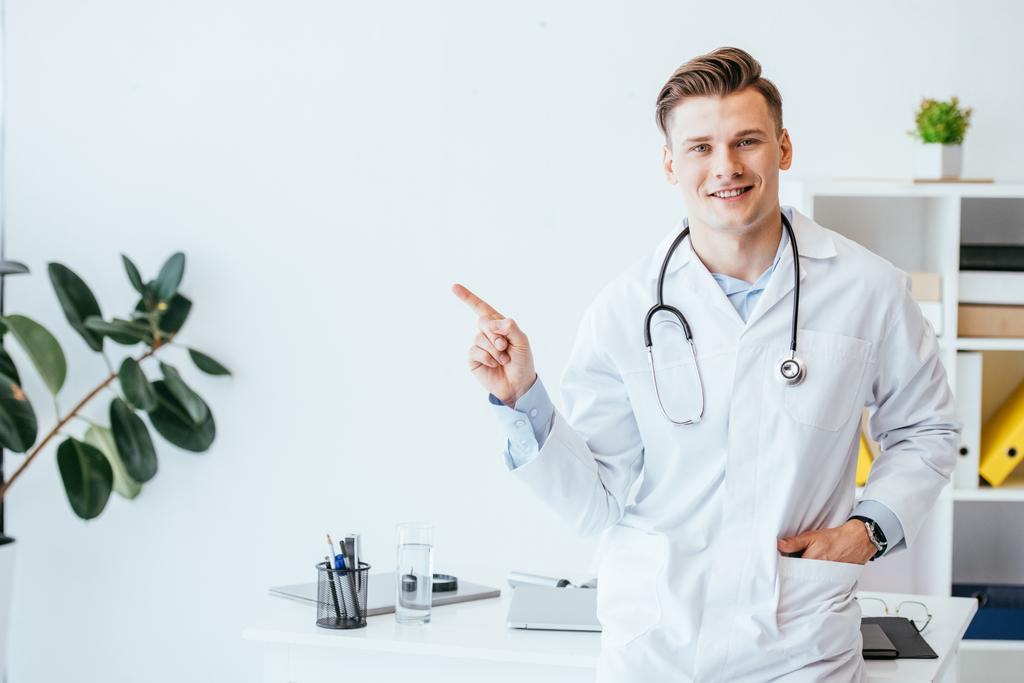 veselý lékař v bílém plášti stojí s rukou v kapse a ukazuje prstem na klinice  - Fotografie, Obrázek
