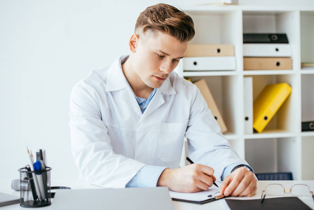 seriöser und gutaussehender Arzt in weißem Kittel schreibt Diagnose auf Klemmbrett  - Foto, Bild