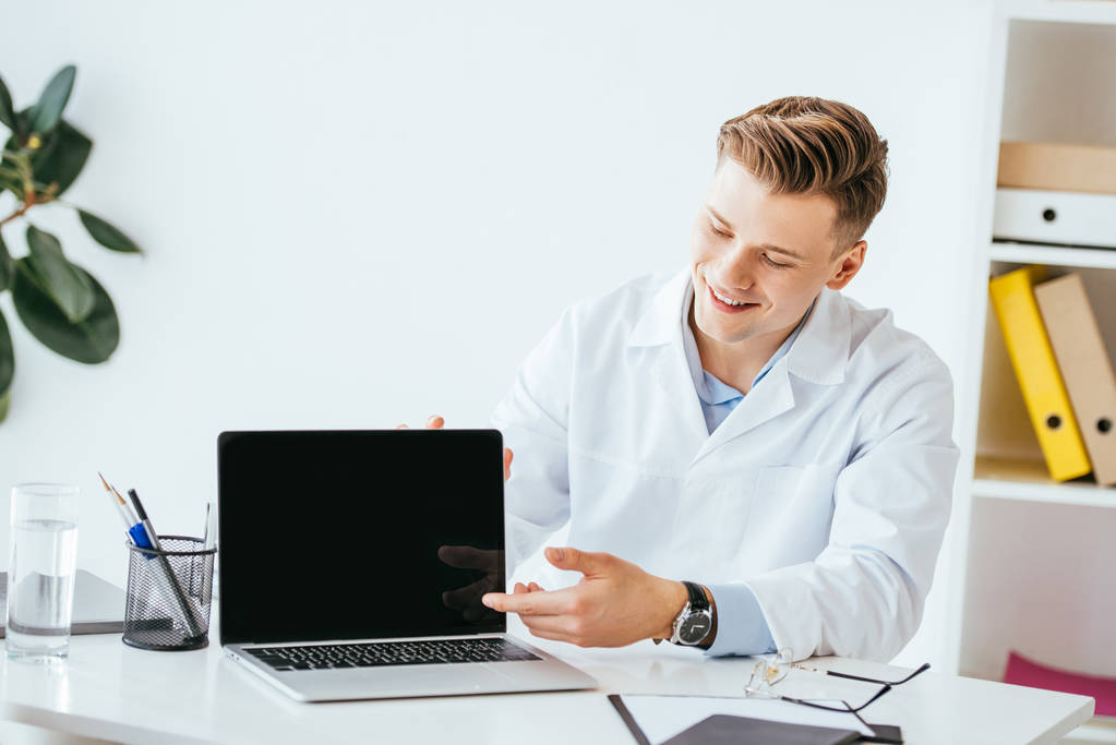 cheerful doctor pointing with finger at laptop with blank screen  - Photo, Image