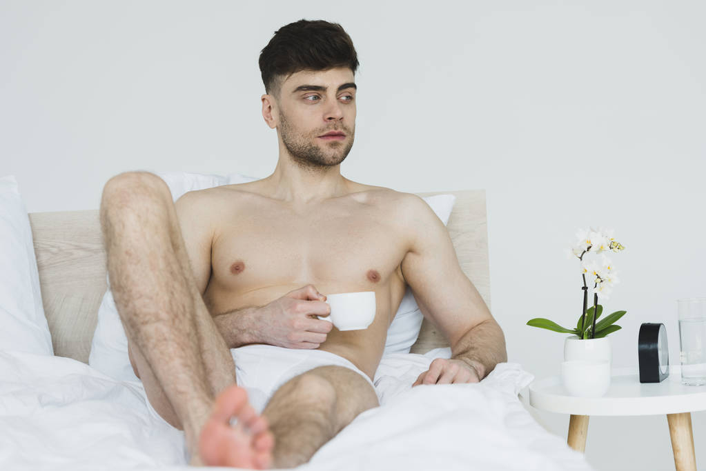 selektywne skupienie przystojnego, przemyślanego mężczyzny w spodniach leżącego w łóżku z filiżanką kawy - Zdjęcie, obraz