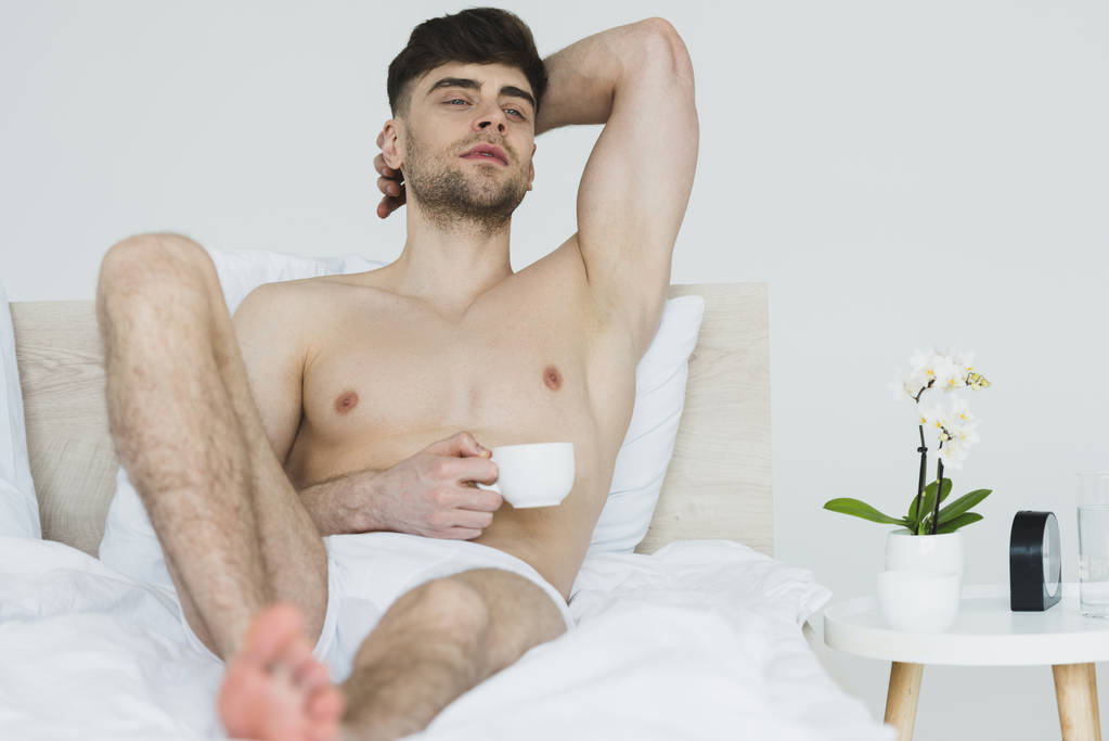 enfoque selectivo de hombre sonriente pensativo en calzoncillos acostado en la cama con taza de café
 - Foto, imagen