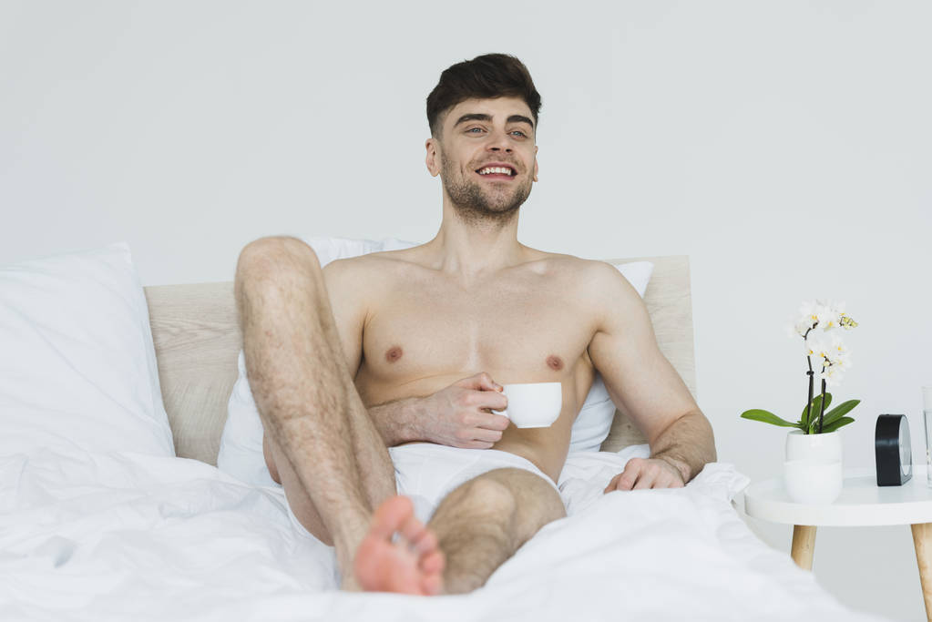 selective focus of handsome smiling man in underpants lying in bed with cup of coffee - Photo, Image