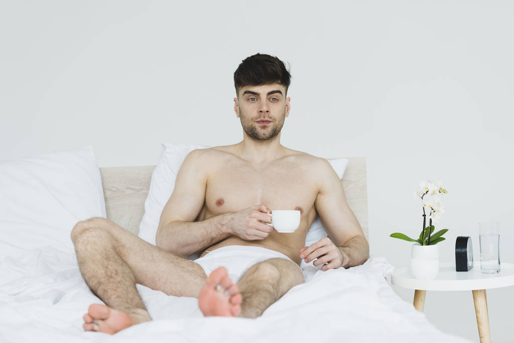 selectieve focus van knappe, doordachte man in onderbroek liggend in bed met kopje koffie - Foto, afbeelding