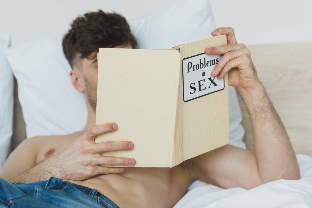 selective focus of shirtless man in blue jeans reading problems in sex book while lying in bed - Photo, Image