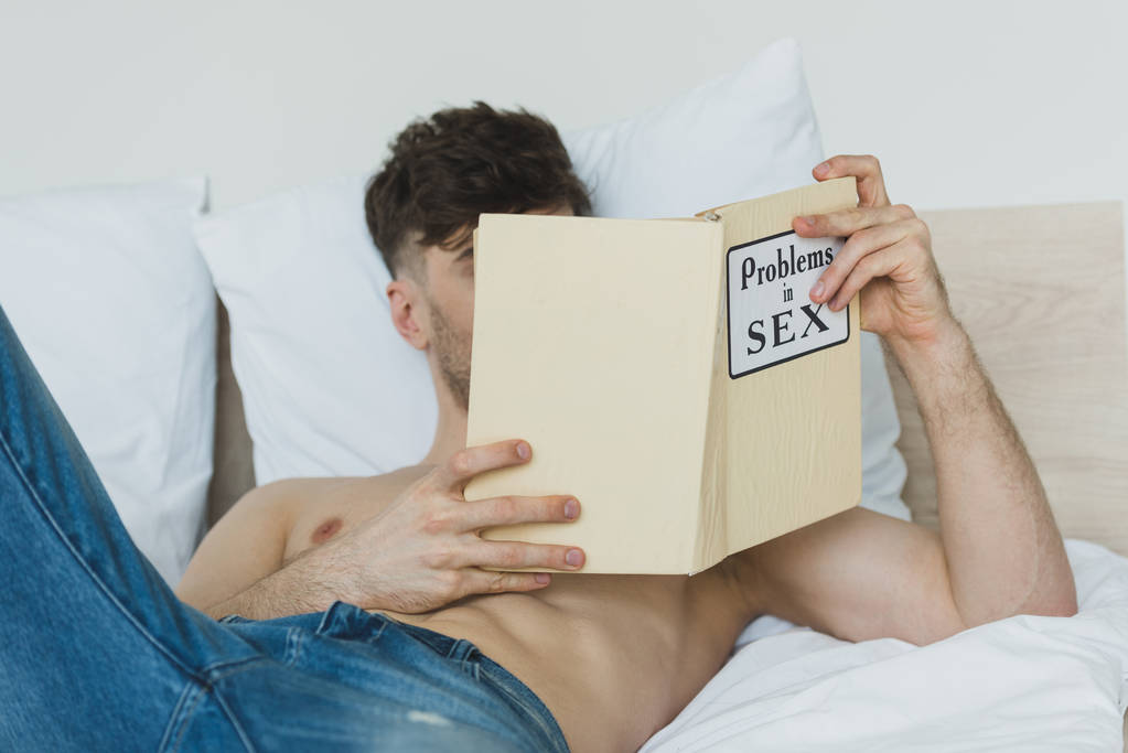 白い寝具の上に横たわっている間、青いジーンズの読書問題のシャツレスの男の選択的な焦点 - 写真・画像