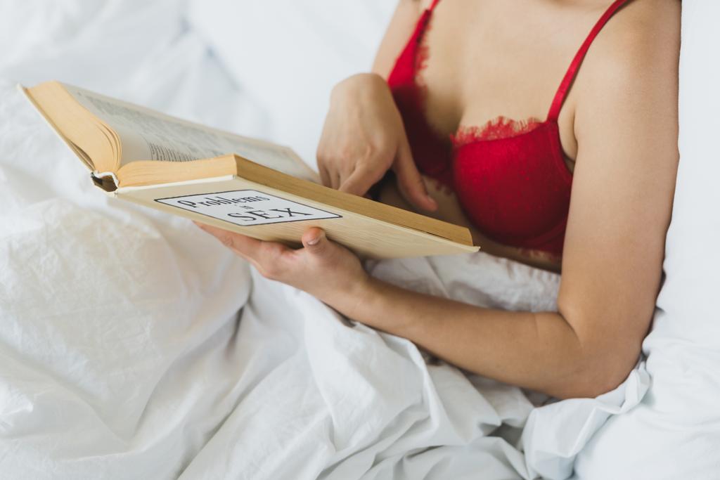περικομμένη όψη της γυναίκας με κόκκινο σουτιέν που βρίσκεται διαβάζοντας προβλήματα στο σεξ βιβλίο, ενώ ξαπλωμένο στο κρεβάτι - Φωτογραφία, εικόνα