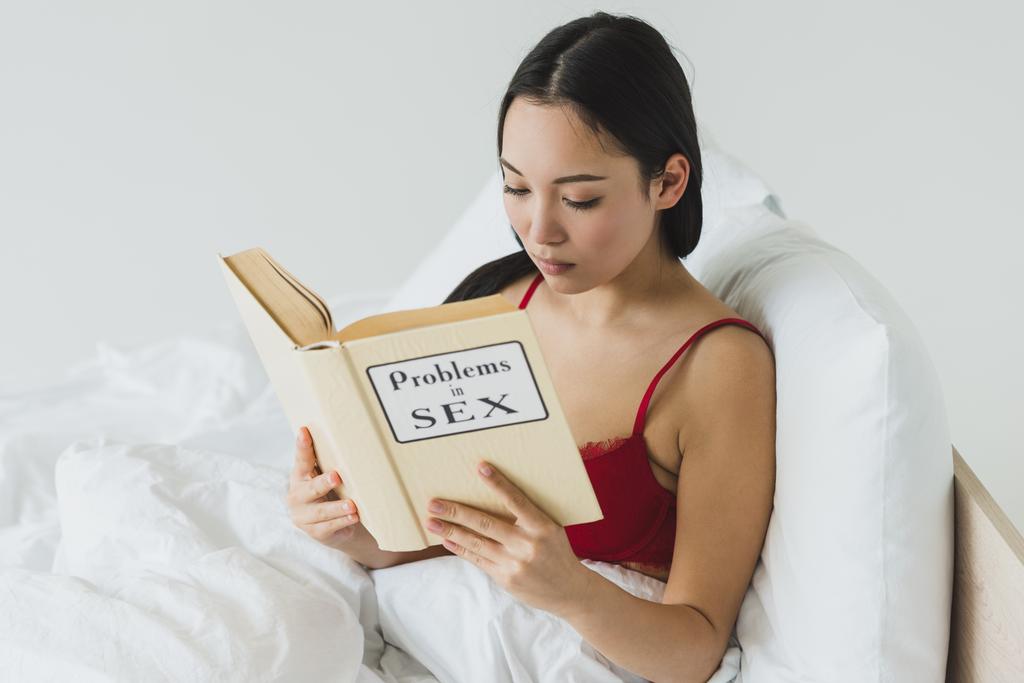 όμορφη Ασιατική γυναίκα στο κόκκινο σουτιέν διαβάζοντας προβλήματα στο σεξ βιβλίο, ενώ ξαπλωμένο στο κρεβάτι - Φωτογραφία, εικόνα