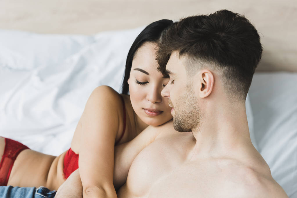 schöne asiatische Frau in roten Dessous umarmt hemdlosen Freund, während sie zusammen im Bett liegt - Foto, Bild