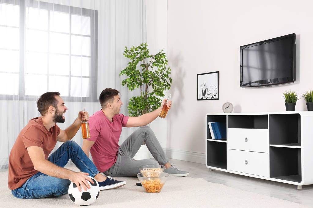 Hombres jóvenes viendo la televisión mientras están sentados en el suelo en casa. Canal deportivo
 - Foto, Imagen