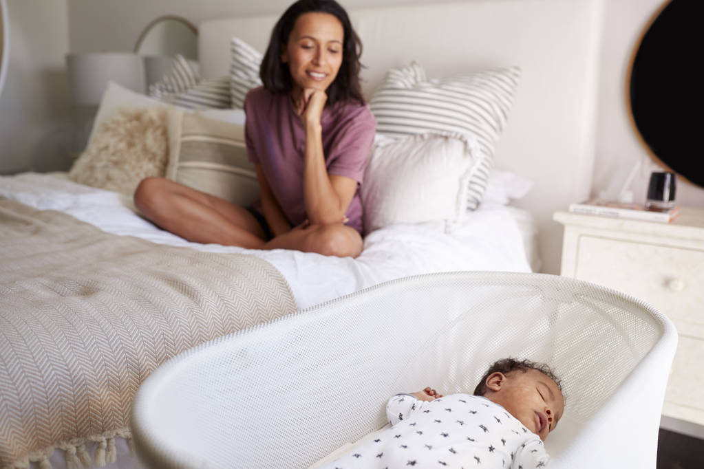 Genç yetişkin anne yatağında bacak bacak üstüne atarak oturur ve üç aylık bebeğinin onun karyolasında uyumasına bakar. - Fotoğraf, Görsel