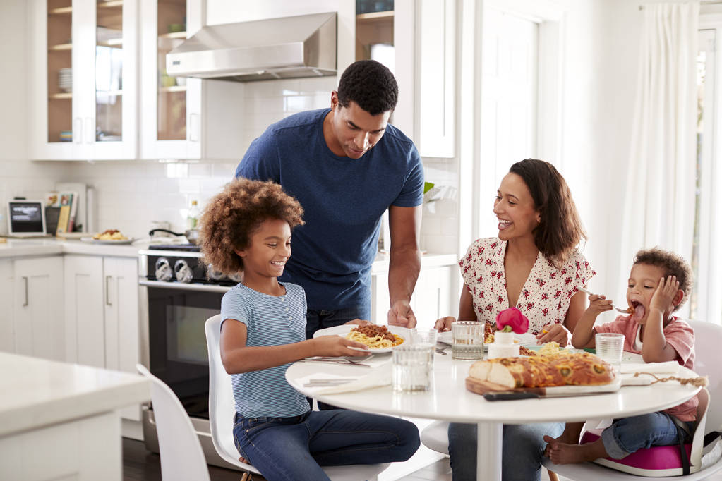 junge Mutter sitzt mit Kindern am Tisch in der Küche, Vater serviert ihnen Essen - Foto, Bild