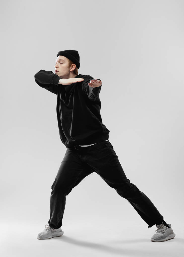 Танцор, одетый в черные джинсы, толстовку, шляпу и серые кроссовки танцует, делая движения руками в студии на белом фоне
 - Фото, изображение