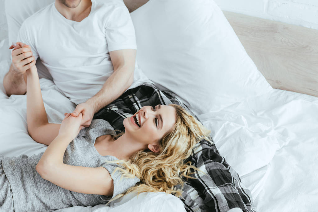обрезанный вид счастливой блондинки, держащейся за руки с парнем, лежащим на кровати
 - Фото, изображение