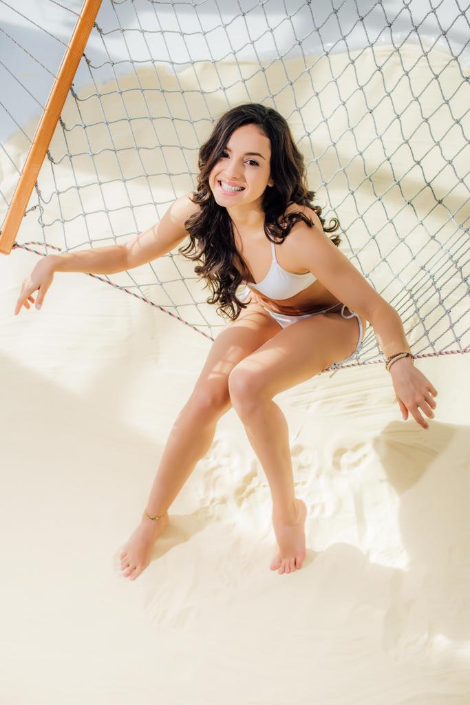 hermosa chica sexy en bikini sonriendo y mirando a la cámara mientras está sentado en hamaca en la playa
 - Foto, Imagen