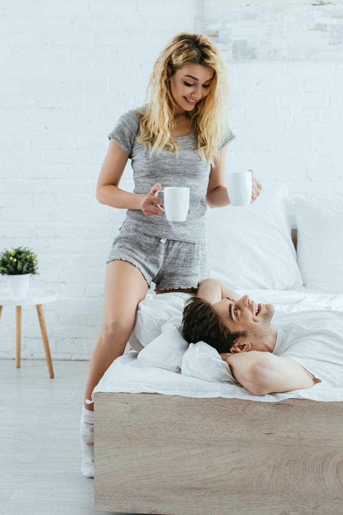 ベッドに横たわっている幸せな男の近くにコーヒーとカップを保持する陽気なブロンドの女性  - 写真・画像