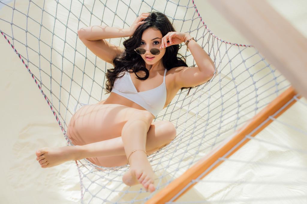 beautiful girl in bikini and sunglasses lying in hammock and looking at camera on beach - Photo, Image