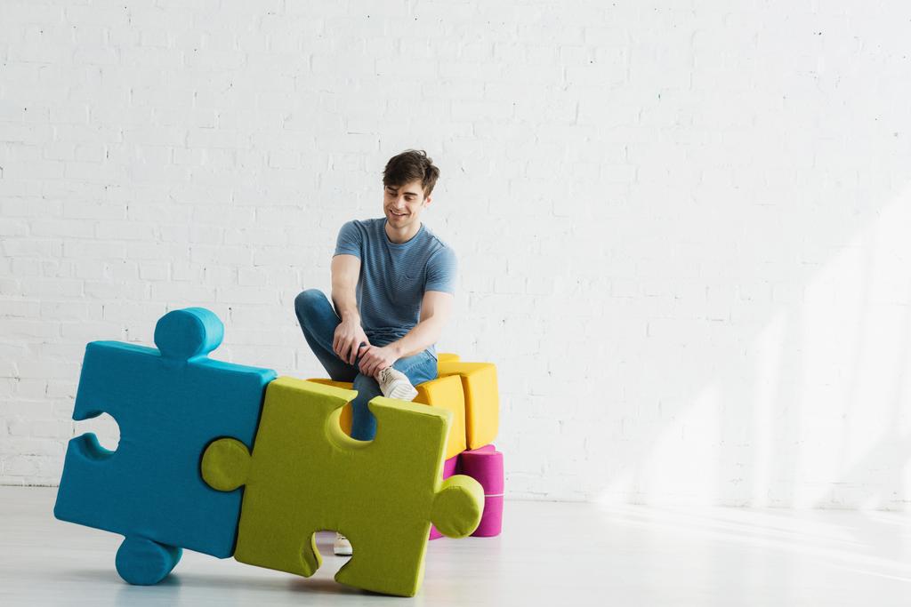 fröhlicher Mann, der blaue und grüne Puzzleteile betrachtet, während er in der Nähe der Ziegelmauer sitzt  - Foto, Bild